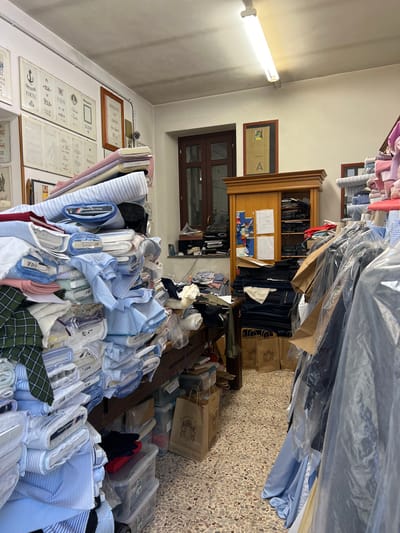 The image depicts a cluttered tailor shop in Ferno, Italy, likely a small business. The scene is dominated by large stacks of fabric rolls, predominantly in blues, whites, and stripes, indicating a focus on shirting materials. Some rolls have labels visible, suggesting they are for sale or use in tailoring. To the left, several framed certificates or awards are mounted on the wall. In the center, a wooden cabinet with sliding doors is filled with more fabric rolls and what appear to be tailoring supplies. A window is visible above the cabinet. To the right, racks of clothing covered in plastic garment bags are present, along with additional stacks of fabric. Several brown paper shopping bags are visible on the floor and near the cabinet, some with printed logos. The overall impression is one of a working tailor's studio, packed with inventory and materials. There are no people visible in the frame.
