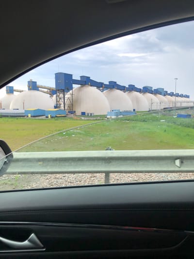 The image displays an industrial scene, likely viewed from inside a vehicle. The prominent subjects are a long row of large, light-colored spherical storage tanks, resembling those used for liquefied natural gas (LNG) or other gases, extending into the distance. Each spherical tank is connected to or accompanied by smaller, blue-colored industrial structures, which appear to house operational equipment, pipework, and support frames. Some of these blue structures feature yellow detailing.
In the mid-ground, a wide, flat expanse of green grass or field separates the viewer from the tanks. There are some lighter, possibly drier or dirt patches within the grass. A grey concrete barrier or guardrail is visible in the foreground, separating the immediate roadside from the field. Beyond this barrier, a gravel-covered area indicates a roadside or shoulder.
The photograph is taken from within a dark-colored vehicle, with parts of the interior, including the window frame and a car door panel with a handle, visible on the left and right sides of the frame. The sky is overcast with light grey clouds. The scene conveys an industrial setting, consistent with the port and energy infrastructure found in Ust-Luga, Russia. No people or discernible text are visible in the image. The time of day is daytime.