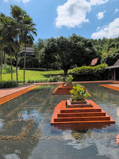This image captures a tranquil outdoor scene at the Raya Heritage hotel in the city of Ban Pong Nuea, Thailand, on a bright, sunny day with scattered white clouds against a blue sky. In the foreground, a shallow, rectangular body of water with a tiled bottom and smooth, dark grey tiles is visible. Within the water, several rectangular, stepped platforms constructed from reddish-orange bricks rise above the surface. Each platform is topped with a planter containing lush green plants with large, round leaves, resembling lotus plants. The water's surface is rippled, reflecting the sky and the surrounding greenery. The edge of the pool and the platforms are lined with the same reddish-orange bricks.

In the mid-ground, beyond the water feature, a meticulously maintained green lawn stretches out, bordered by tall, lush green grasses and reeds. Several slender palm trees with fronds reaching towards the sky are positioned to the left of the lawn. Behind the lawn and palm trees, a multi-story structure with a dark roof and balconies is partially visible, nestled amongst dense foliage. Further back, a large, mature tree with a broad canopy dominates the center of the frame, and a traditional-looking building with a terracotta tiled roof and dark wooden eaves is situated to the right, surrounded by dense, manicured hedges. The background is filled with the verdant slopes of a hill covered in a variety of trees and vegetation, suggesting a natural, forested environment. The overall impression is one of serene natural beauty and refined tropical architecture.