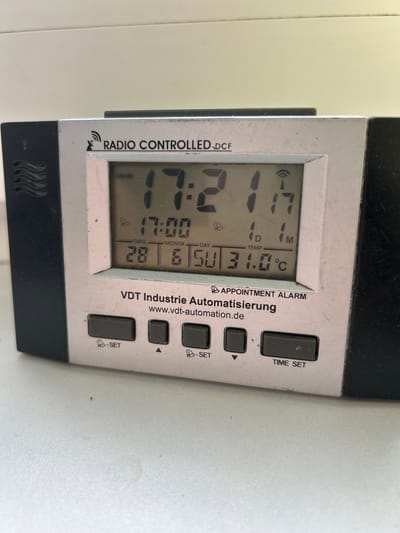 A digital clock radio with a silver and black casing is displayed on a white surface. The clock face shows the time as 17:21 and the date as 28.6.50. The temperature is displayed as 31.0°C. The brand name "VDT Industrie Automatisierung" is visible below the display. The clock is radio-controlled, indicated by the text "RADIO CONTROLLED - DCF". Several buttons are located below the display, labeled "SET", an upward-pointing arrow, and "TIME SET". The environment appears to be indoors, with a slightly textured white surface beneath the clock. There are no people or animals present in the image. The image does not contain any information that would allow for determining its location in Teplice, Czechia.