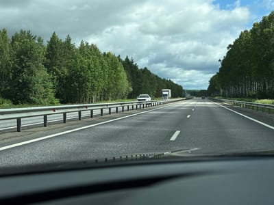 The image depicts a view from inside a vehicle traveling on a multi-lane highway flanked by dense green forests on both sides. The highway has clear white lane markings. In the distance, there are several vehicles, including a white truck with a white trailer and a white SUV, and a darker colored car further down the road. The sky is cloudy with patches of blue. The time appears to be daytime, likely afternoon, given the lighting. The location is specified as Pologovka, Russia.