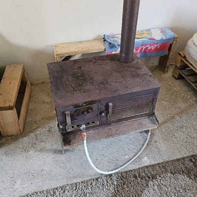 An interior photograph taken in the city of Buriani, Georgia, depicts a rustic room dominated by a dark, rectangular metal wood-burning stove exhibiting surface rust, situated on a concrete floor. A brown, shaggy rug partially covers the foreground. A dark-colored stovepipe extends vertically from the top of the stove. A white flexible hose is connected to a brass fitting on the front lower section of the stove, with some reddish discoloration visible around the connection point on the stove. To the left of the stove, an open, unpainted wooden crate rests on the concrete. In the background, against a light-colored wall, a simple wooden bench is present, draped with a blue and white towel featuring red lettering. To the far right, a stack of wooden pallets supports a white object, possibly a mattress or cushion. No individuals are present in the scene.