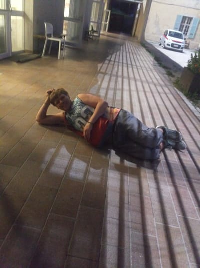 A young person with short, brown hair lies on their left side on a tiled floor outdoors. They are wearing a sleeveless, dark tank top with a graphic design in blue and white, and a red undershirt. Their right arm is bent with their hand resting on their head, and their left arm is resting on their hip. They are wearing dark pants and dark shoes. The tiled floor has a pattern of light and dark rectangular tiles, and shadows from vertical bars are cast across it. In the background, to the left, is a building with large windows and a white chair. To the right, a white car with a red stripe is parked on a paved area. The overall scene appears to be in a public area, possibly a courtyard or walkway, in Levico Terme, Italy.