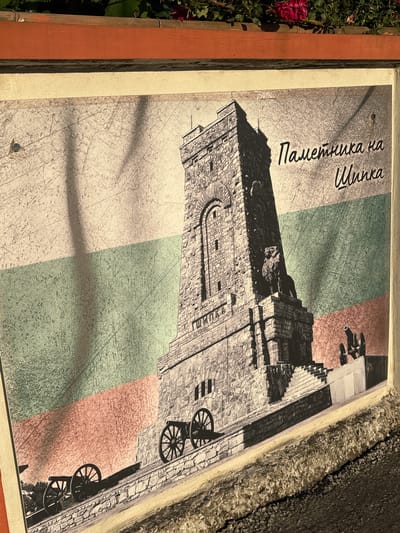 The image displays a monument in the city of Enevo, Bulgaria, against the backdrop of the Bulgarian national flag. The monument, rendered in a sepia tone, is a stone tower with intricate carvings and features a lion statue at its base. Cannons are positioned on either side of the monument, suggesting a historical or military context. The flag's white, green, and red stripes are visible behind the monument, with diagonal shadows cast across the scene. The overall composition is reminiscent of a vintage postcard or historical photograph. The text on the image reads "Паметника на Шипка," which translates to "Monument to Shipka."