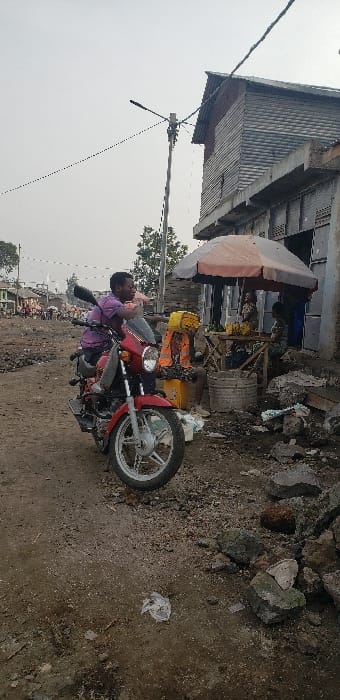 The scene depicts an unpaved street in Buzove, Democratic Republic of the Congo. In the foreground, a red motorcycle, resembling a Promax model, is positioned on a dirt and gravel road. A male individual, wearing a purple collared shirt, is seated on the motorcycle, looking towards the right side of the frame. To the right of the motorcycle, a vendor's stall is established under a light-colored umbrella. The stall displays bananas and other produce on a wooden table. An individual wearing an orange high-visibility vest is partially visible near the stall, and another person is seated behind it. The ground in the immediate vicinity is uneven, comprising dirt, loose rocks, and scattered refuse, including a clear plastic bag. A multi-armed utility pole with overhead power lines stands centrally. In the background, a two-story structure with corrugated metal siding and concrete or brick elements is visible. Further into the distance, additional buildings and indistinct human figures suggest a populated area under an overcast sky.