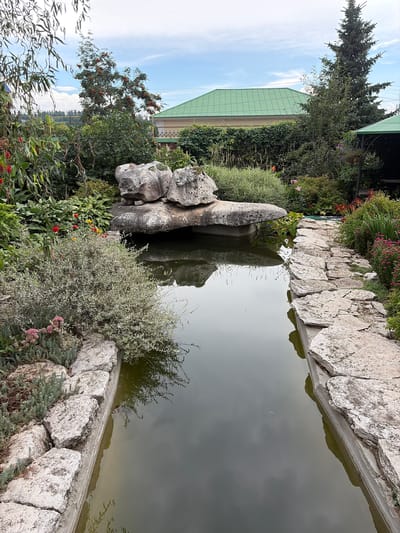 This is a medium-shot, eye-level, vertical, outdoor, daytime, full color photograph of a garden pond in Norja, Russia. The pond is rectangular and lined with large, rough-cut stones. The water is murky and reflects the sky and surrounding foliage. In the center of the pond, a large, irregularly shaped rock formation sits precariously on a narrower stone. Lush green plants and trees surround the pond, with some red and yellow flowers visible on the left side. In the background, a house with a green roof and some electrical wires are visible, along with a tall evergreen tree on the right. The sky is partly cloudy. There are no people or animals visible in the image.