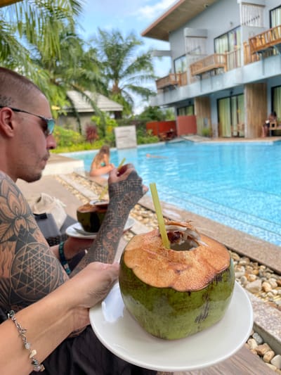 A male individual with short dark hair, wearing reflective blue sunglasses and extensive geometric tattoos on both arms and shoulder, is seated in the foreground, looking to his right. He holds a green coconut drink with a yellow straw on a white plate in his left hand, and another similar coconut drink in his right hand. He wears a dark garment and a light-colored beaded bracelet on his left wrist. In the mid-ground, a clear blue swimming pool is visible. A person with light-colored hair, wearing a blue swimsuit, is sitting at the pool's edge, partially submerged, facing away from the camera. Another person is visible swimming further in the pool. The background features a multi-story light-grey building with wooden balconies, indicative of a resort, and dense tropical foliage, including numerous palm trees, under a bright, partly cloudy sky. The scene is located in Ban Tha Muang, Thailand.