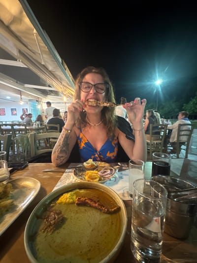 A woman with curly brown hair and glasses is eating a skewer of food at a restaurant in Asprovalta, Greece. She is wearing a blue and orange floral dress and has tattoos on her arm. The restaurant appears to be outdoors with tables and chairs set up. There are other people in the background dining. On the table in front of the woman are plates of food, drinks, and cutlery. The lighting suggests it is nighttime.