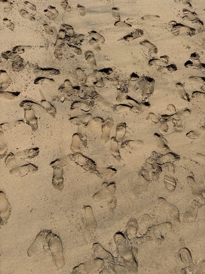 The image depicts a sandy surface, likely a beach, with numerous human footprints scattered across it. The sand is a light tan color and appears to be slightly damp. The footprints vary in size and depth, suggesting that different people or the same person at different times have walked across the area. The scene is located on a beach in Pájara, Spain. There is no other human presence, other animals, or objects visible other than the sand and footprints.