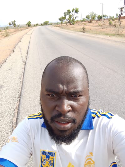 A close-up selfie of a man with dark skin, a shaved head, and a black beard is taken on a paved road. He is wearing a white soccer jersey with blue and yellow stripes and patches. The road stretches into the distance, curving slightly to the left. The surroundings consist of dry, arid land with sparse vegetation and scattered trees. In the far background, there are buildings, possibly a village or small town. The photo seems to be taken on the outskirts of a populated area, possibly near Mulke Barikin Sayejo, Nigeria.