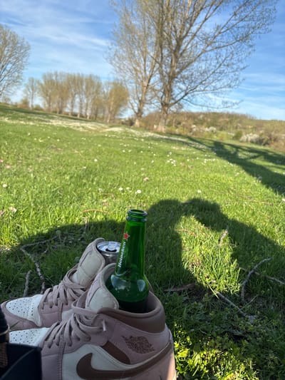 The image depicts a grassy field under a partly cloudy sky, likely in Beli Brod, Bulgaria, though this is not explicitly confirmed in the image. In the foreground, a pair of light-colored sneakers sit on the grass, with a green Heineken beer bottle positioned between them. The bottle appears to be empty. The background consists of trees and more grassy area, typical of a park or rural landscape. The overall impression is one of leisure and relaxation.