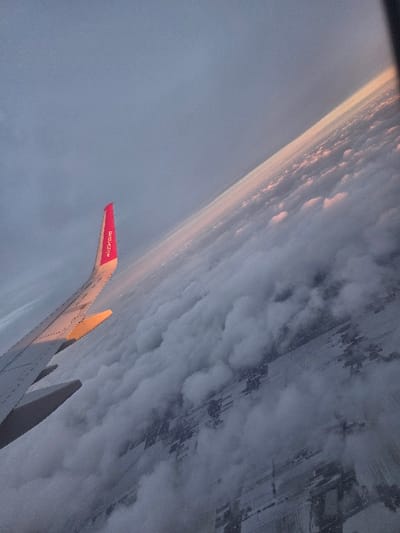 A tilted aerial photograph depicts a landscape over or near Bronisze, Poland, captured from an aircraft window during low-angle sunlight conditions, consistent with dawn or dusk. The lower left portion of the frame is occupied by a gray aircraft wing, its upper surface partially illuminated by golden light. A pink winglet, displaying the text "WIZZ AIR", extends upwards from the wing's trailing edge. Below the aircraft, an extensive, broken layer of white and gray clouds partially obscures the ground. Through gaps in the cloud cover, snow-covered terrain is visible, characterized by discernible rectangular field patterns and faint indications of scattered structures. The upper section of the image shows a blue-gray sky, which transitions to a horizontal band of soft pink and orange illumination at the distant horizon.