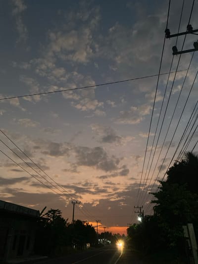 The image captures a twilight scene along a paved road in Ierhobglumpang, Indonesia. The sky displays an upper section of blue-grey with scattered light-colored cumulus clouds, and a faint, bright celestial body is visible in the upper left quadrant. Towards the horizon, the sky transitions to warm orange, pink, and yellow hues. Multiple dark electrical power lines crisscross the frame from foreground to midground, connecting to utility poles visible on both sides of the road. On the left, the dark silhouette of a low-rise building, possibly residential, with recessed window openings, is visible, flanked by dark trees and shrubs. On the right, a dense mass of dark green foliage, including a prominent palm frond in the upper right, borders the road. The paved road recedes centrally into the distance, where the bright, undiffused headlights of an approaching vehicle are prominently visible. Several other faint artificial lights appear further down the road beyond the vehicle. No individuals are present in the scene.