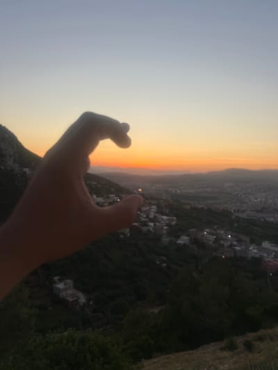 This vertical image, captured in Tétouan, Morocco, showcases a panoramic view of a city at sunset or dusk from an elevated vantage point. A human hand, silhouetted against the bright sky, is prominently featured in the lower center-right foreground. The hand is cupped with the thumb extended upwards, seemingly framing or holding the distant sun.

The sky displays a striking gradient, transitioning from a pale grey-blue at the top to warm orange, yellow, and reddish hues at the horizon, indicating either sunset or sunrise. The weather appears clear, with no clouds visible. Below the horizon, a sprawling city or town is nestled within a valley or along hillsides. Numerous buildings are visible, some with distinct lights beginning to twinkle, confirming the evening hour. The surrounding terrain is rugged and mountainous, covered in dark vegetation. The overall scene conveys a serene and expansive view of the urban landscape meeting the natural environment under a dramatic sky. No text is visible in the image.