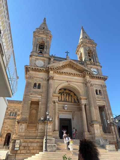 This is a low-angle, vertically oriented photograph of the facade of a stone church, likely a cathedral, in Alberobello, Italy. The clear blue sky indicates it is daytime, with the sun casting shadows and highlighting the textured stone architecture. The church features two prominent towers, one on the left with a spire and bells visible, and another on the right with a large clock face. The main entrance is set within a grand arched portico, adorned with ornate carvings and sculptures. Several people are visible in front of the entrance, some appearing to be entering or exiting the building, while others are gathered on the steps.

Notable architectural details include intricate stonework, decorative columns, and a large sun dial on the tower. The overall impression is of a historical and significant religious structure. The presence of tourists suggests it is an active tourist site. There is a sign with text and images near the entrance, but the text is too small to read. The lighting and bright sky suggest it is a sunny day, likely in the late morning or early afternoon.
