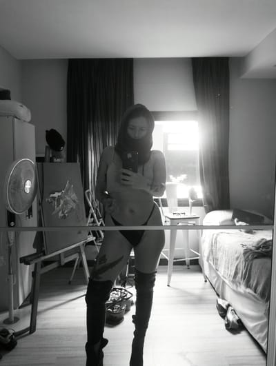 A black and white image depicts a person taking a selfie in a mirror within a domestic interior. The individual is wearing thigh-high boots, a thong, and a crop top, with their face obscured by a garment pulled up over their head. Visible tattoos adorn their arms and legs.
The room contains an easel with an unfinished painting, an electric fan, a bed with rumpled linens, a white stool with glasses and a wine glass, and various containers. The setting appears to be a bedroom or studio. The location in Inriville, Argentina is not determinable from the image content.