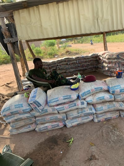 A man in military attire is seated beside a stack of cement bags branded "Dangote Cement" within a simple, open-sided structure. The structure, partially constructed with wooden posts and corrugated metal, offers a view of a rural landscape. Sandbags line the perimeter, providing a barrier or fortification. The bags of cement are stacked high, possibly for construction or storage purposes. The scene evokes a sense of a security post or construction site in a developing area.