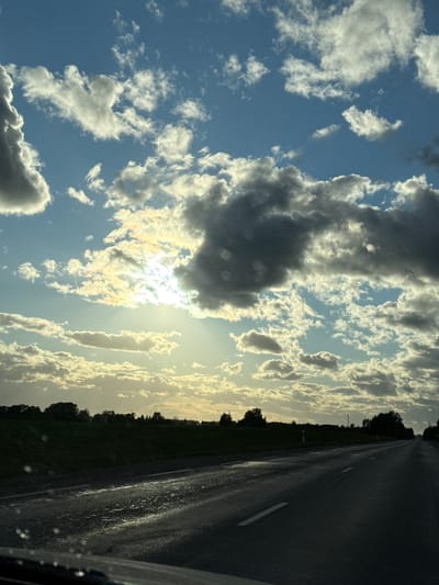 The image shows a view from a vehicle on a road. To the right of the frame is a wet asphalt road with a white line marking a lane. Beyond the road is a line of trees and foliage silhouetted against the bright sky. The majority of the frame is occupied by the sky, which is partially obscured by clouds. The sun is shining through a gap in the clouds, creating a bright glare. The sky is blue in areas with thinner clouds, contrasting with the darker grey clouds. There are no people visible in the image. Although the image is taken on a road in Beržiniai, Lithuania, the road name or any objects that would show that are not visible.
