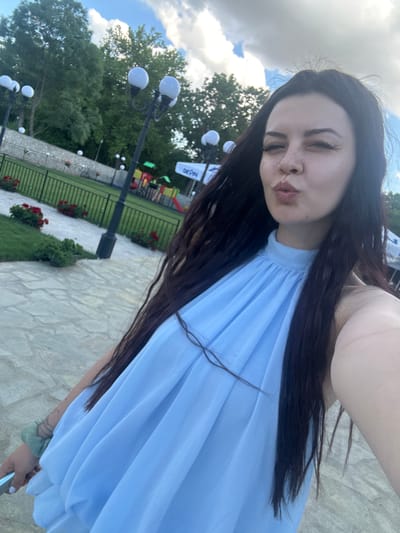 A young woman with long, dark hair is posing for a selfie outdoors. She is wearing a light blue, sleeveless dress with a high neckline and a ruffled bodice. Her lips are pursed as if blowing a kiss, and she is winking. The background shows a park-like setting with trees, lampposts, and a playground with colorful slides and swings. A white sign with the word "DEVIN" is visible in the distance. The ground is paved with light-colored stones. The image appears to have been taken during daylight hours, with a cloudy sky. The location is the city of Zlati Voyvoda, Bulgaria.