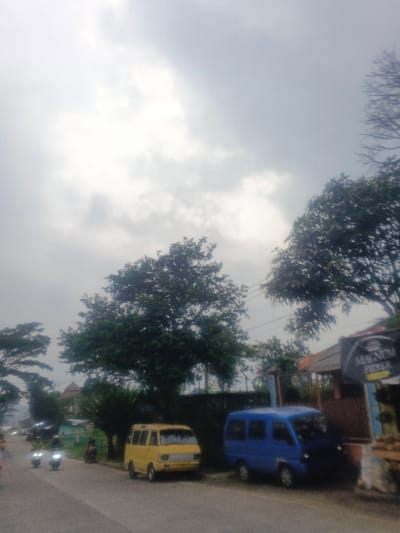 The image displays a daytime street scene in the city of Morrumbala, Mozambique, under a bright, overcast sky. A paved road runs diagonally from the lower-left foreground into the upper-right distance. Two small vans are parked on the right side of the road: a yellow van is positioned closer to the viewer, facing left, while a blue van is parked immediately behind it, adjacent to a building. In the lower-left portion of the road, two motorcycles are in motion, each operated by an indiscernible individual, with their headlights illuminated as they travel away from the viewer. On the right side, a building with a dark brown facade and a tiled roof is partially visible, featuring a sign displaying the word "MAGNUM". Multiple trees with dense green foliage are present throughout the scene, including a large tree in the central-left and another partially obscuring the building on the right. The environment is consistent with a semi-urban setting.