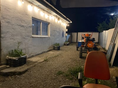 The image depicts an outdoor scene at night, illuminated by a string of overhead lights. In the foreground, the back of an orange chair is visible, suggesting a seating area. Behind the chair, a small orange and black tractor or tiller is parked on a gravelly surface. The tractor has large wheels and appears to be an agricultural or gardening implement.
To the left of the tractor, a white building with a textured wall and a window is visible. A string of incandescent bulbs hangs along the roofline of the building, providing a warm glow. Planters filled with greenery are placed near the building.
In the background, a corrugated metal fence separates the property from what appears to be more open land or other structures. Trees are visible beyond the fence, and a sliver of the moon is visible in the dark night sky. The overall setting suggests a rural or semi-rural location, possibly a backyard or farmstead in Kazakhstan, given the location context. There is no visible text in the image. The scene appears calm and quiet, with the artificial lighting creating a somewhat cozy atmosphere despite the darkness.