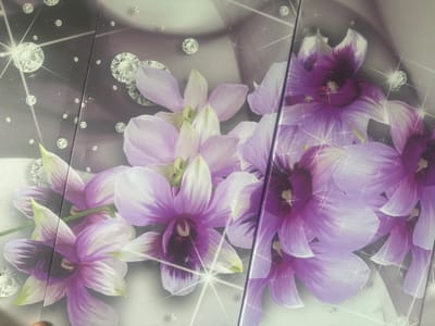 The image shows a close-up view of a decorative panel featuring purple orchids and sparkling diamonds. The panel is divided vertically in the center, suggesting it is part of a larger surface like a wall or furniture. The orchids have delicate petals with varying shades of purple and white, some with dark purple centers. The diamonds are depicted as faceted, sparkling gems scattered around the flowers, adding a touch of glamour. The background is a soft gradient of grey and white, creating a gentle contrast with the vibrant flowers. The overall impression is one of elegance and luxury. There is no information in the image that indicates its location in Stepnogorsk city administration, Kazakhstan.