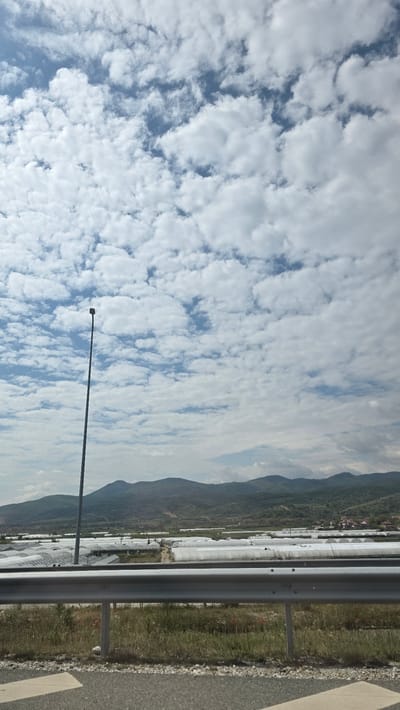 The image, likely taken from a moving vehicle, shows a view towards a field of greenhouses near Mikrevo, Bulgaria. A highway guardrail is visible in the foreground, with a stretch of paved road adjacent. Beyond the guardrail lies vegetation, with a long expanse of greenhouses extending towards the background. Mountains form the backdrop. The sky is filled with numerous small, white clouds against a blue background, and a streetlight pole stretches diagonally across the lower portion of the frame.
