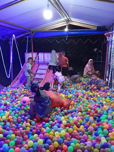 The image depicts a children's ball pit play area in Namplohbaro, Indonesia, illuminated by artificial light, suggesting evening or night. The facility is covered by a corrugated metal roof supported by metal framing, with blue fabric draped along the upper perimeter. The play area is enclosed by a dark chain-link fence or netting, visible in the background. Overhead, a bright spherical light bulb hangs from the roof, and purple LED strip lights are installed along the structural framing and possibly around a basketball hoop structure on the right.
The entire ground surface is filled with a dense collection of multi-colored plastic balls. A central children's play structure, featuring a pink slide and stairs, is situated within the ball pit.
Numerous individuals are present:
* **Children:** Approximately seven children are visible engaging in play. One child in light-colored, patterned clothing is descending the pink slide. Another child, wearing a light green headscarf (hijab) and light top, is climbing the stairs of the play structure. A child in patterned pajamas is at the base of the slide. Another child in a dark top and light pants kneels in the ball pit near the slide. A child wearing a plaid shirt is seated in the ball pit, holding a small red and white ball. One child, with their back to the viewer and dark hair tied in pigtails, is seated near a large orange inflatable object. A child in a red shirt is partially visible behind the stairs.
* **Adults:** At least one adult woman is seated on the edge of the ball pit on the right, wearing a light pink headscarf (hijab) and a patterned garment, looking towards the camera with a slight smile. Another adult is partially visible behind the stairs.
The scene conveys active children's recreation within a brightly lit, contained play environment.