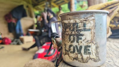 A close-up shot focuses on a weathered metal mug with a sticker on its side. The sticker is partially torn and smudged, but the words "LOVE" are clearly visible in bold, dark lettering. Above "LOVE," remnants of another word or phrase are present, but illegible. The mug has a carabiner attached to its handle, which is wrapped with black cord.

In the background, a blurred scene depicts an outdoor setting. A person with dreadlocks is visible, wearing dark clothing and a golden necklace. Other individuals and objects are out of focus, suggesting a gathering or a temporary living space. Red and blue fabrics are spread out, and a cardboard box is placed on the ground. The overall atmosphere appears casual and perhaps rustic, given the natural lighting and the surrounding environment which hints at a natural or semi-developed location. The lighting suggests it is daytime. The provided location context is Shila, India.