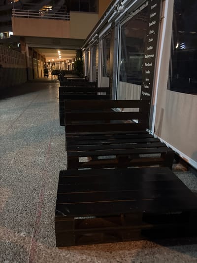 The image shows an outdoor seating area at night, likely in the city of Calp, Spain, with a menu board visible on a restaurant's exterior. The seating is constructed from dark wooden pallets and includes benches and tables. The ground is paved with textured concrete. In the background, a narrow walkway leads into a covered area with lighting. Two people are faintly visible walking in the distance, one pushing a stroller. The overall atmosphere suggests a quiet evening at a casual dining establishment.