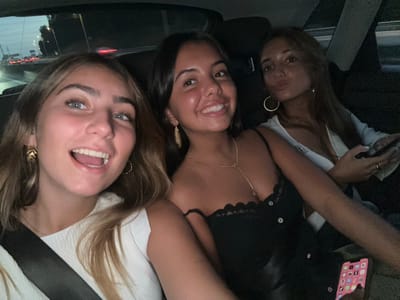 The image displays three young adult females seated across the back of a vehicle, located in Ermesinde, Portugal, during nighttime or dusk. The individual on the left has light brown hair and blue eyes, exhibiting an open-mouthed smile while wearing a white textured top, gold-toned leaf-shaped earrings, and a visible dark seatbelt. The central individual has dark hair and eyes, a closed-mouth smile, and wears a black lace-trimmed top with front buttons, a gold-toned necklace, and gold-toned drop earrings. The individual on the right has light brown hair, poses with puckered lips, and wears a white top with a gold-toned hoop earring, holding a black smartphone in her left hand. A second smartphone, displaying a grid of colorful application icons, is partially visible in the bottom right foreground. The background, viewed through the vehicle's rear window, shows blurred lights indicative of a roadway at night.