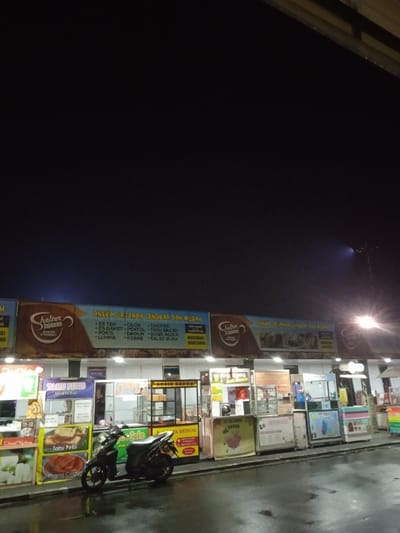 This nighttime image depicts a row of illuminated food stalls, likely a street food market or food court, in Kecamatan Kartasura, Indonesia. The sky above is dark, indicating late evening or night, and the wet, reflective ground suggests recent rain.
The scene is dominated by several vendor kiosks arranged under a long, brightly lit overhead banner. The banner prominently features a "Shelter Jajanan" logo and advertises "ANEKA JAJANAN LENGKAP DAN MURAH" (Variety of Complete and Cheap Snacks). The listed items include "ES TEH," "CILOK," "TAKOYAKI," "ES DAWET," "PENTOL," "TAHU BAKSO," "POKIS," "DIMSUM," "SOSIS BEDUG," "LUMPIA," "KEBAB," and "SALAD BUAH."
Individual stalls below the main banner showcase their offerings. On the left, one stall advertises "TAHU PETIS" (Tofu with shrimp paste sauce) from "Mak Ni," and "Pisang Crispy" (Crispy Banana). A black motorcycle is parked in front of these stalls. Further to the right, other kiosks are visible with signs such as "ES BEDUG" and "MY DAY." Another partially visible sign likely says "KEBAB." The stalls vary in design, some featuring glass display cases and others with colorful graphics.
No people are clearly visible in the image, contributing to a quiet atmosphere despite the vibrant artificial lighting. The bright lights from the stalls and a distant streetlamp on the far right cut through the darkness, illuminating the food vendor area, which appears ready for business.
