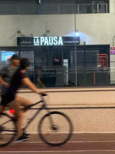 The image depicts a motion-blurred scene of two people on bicycles passing by a restaurant and cafeteria named "LA PAUSA restaurante cafeteria" in Castelló De La Plana, Spain. The sign for the establishment is clearly visible with illuminated white lettering. The building appears to be modern, with large glass windows reflecting the surroundings. Inside the establishment, a red fire extinguisher mounted on a wall is partially visible through the glass. The foreground shows a textured, light-colored tiled wall, and below that, a reddish-brown paved surface, suggesting an outdoor urban setting. The lighting indicates it is evening or nighttime, with artificial lights illuminating the sign and a soft glow emanating from behind it. One cyclist is in the foreground, moving from left to right, with their legs and bicycle wheels significantly blurred due to motion. Another person on a bicycle is behind and to the left, also blurred. A screen displaying "CRISPY CHICKEN" is visible through the window on the left side of the building.