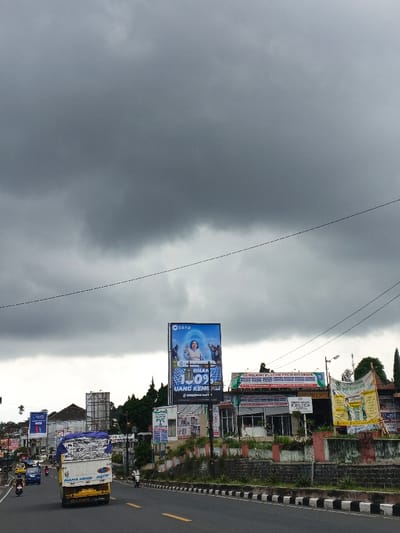 The scene depicts a street in Palayan, Philippines, under a uniformly dark gray, heavily overcast sky. A multi-lane asphalt road with a solid yellow centerline and a black-and-white striped curb barrier on the right dominates the lower half of the image. A white and yellow box truck, equipped with a blue tarp over its cargo bed, is in motion, heading away from the viewer in the right lane. Behind it, a yellow tricycle and a blue sedan are observed in the left lane. A lone motorcyclist operates a dark motorcycle on the far left, traveling towards the viewer. Another motorcyclist is visible on the right shoulder of the road, adjacent to the curb barrier. The right side of the street features a row of commercial buildings, set back from the road by a low, dark-colored wall with red accents and interspersed vegetation. A large, illuminated blue billboard featuring a woman and promoting 'DANA' with text reading 'MENANG 109% UANG KEMBALI' is centrally positioned among these structures. Further signs on the buildings advertise 'Gembilang Plafon PVC & Interior' and display a yellow banner with green graphics. On the left side of the street, further in the distance, additional buildings, another billboard, and traffic light poles are visible. Multiple thin power lines are horizontally stretched across the upper sky.