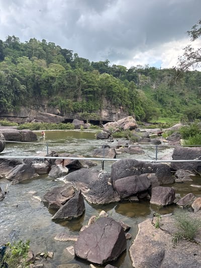 The image depicts a rocky river flowing through a lush, green environment. In the foreground, large, dark boulders are partially submerged in the water, with some emerging to create a rugged riverbed. A metallic railing runs horizontally across the middle ground, separating the foreground from the river beyond. In the middle ground, a person wearing a red shirt is visible wading in the water, holding what appears to be a fishing net. The river expands outwards, surrounded by more rocks and vegetation. In the background, a steep, heavily forested cliff face rises dramatically, with trees clinging to its slopes and rocky outcrops. The sky above is overcast, with dramatic clouds. The scene is located in the vicinity of a waterfall within Tân Phú, Vietnam, likely a natural tourist attraction.
