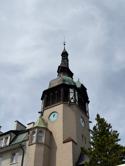 The image shows the exterior of the Sanatorium Lwigród in Świeradów-Zdrój, Poland, focusing on its prominent clock tower. The tower is constructed of light-colored brick and features a dark, ornate spire with a weather vane. Two clock faces are visible on the tower; one is light blue with black Roman numerals, and the other is white with black Arabic numerals. The clock hands on the blue face indicate approximately 12:45, while the white face shows approximately 12:40. The tower has decorative dark wood detailing and arched windows. To the left of the tower, a section of another building with a green, sloped roof and several windows is visible. A large evergreen tree with dense foliage partially obscures the right side of the tower. The sky is overcast with light gray clouds.