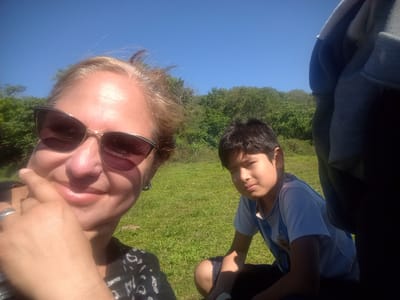 A woman and a boy are in a grassy area under a clear blue sky. The woman is taking a selfie, wearing sunglasses and a patterned top. She is touching her face with her hand, which has a ring on one finger. The boy is sitting on the grass, wearing a blue and white striped soccer jersey. The background includes trees and possibly shrubs or bushes. While there is no definitive visual evidence confirming the exact location, the picture suggests a park or green space in Corrientes, Argentina.