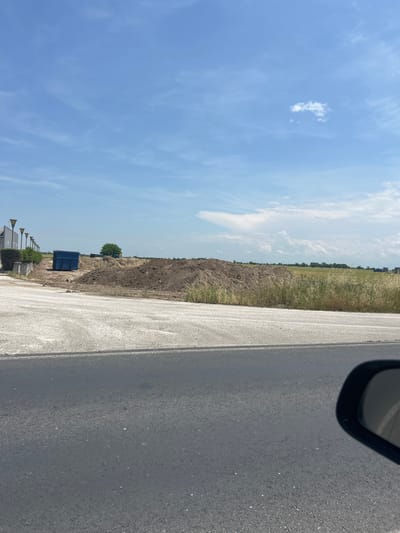 The image displays a sunny, daytime scene in Graf Ignatievo, Bulgaria, captured from a moving vehicle. The primary subjects include an asphalt road, an adjacent construction or land-clearing area, fields, and a vast blue sky.
In the immediate foreground, a dark asphalt road with a lighter-colored concrete or gravel shoulder is visible, extending from the bottom to the top right of the frame. The curved edge of a car's rearview mirror partially obstructs the upper right corner. To the left of the road, a substantial mound of dark brown, excavated soil dominates the midground. Beyond this dirt pile, a field with tall, dry grass and some greener vegetation stretches towards the horizon.
Notable objects within the scene include a prominent dark blue dumpster situated near the dirt mound and a single, lush green tree standing further back. A small, yellow piece of construction equipment, possibly an excavator or loader, is partially visible behind the dirt pile. In the distant background, faint structures and additional fields can be discerned under a bright blue sky adorned with scattered white cumulus clouds.
The weather is clear and sunny, indicative of midday or early afternoon. No people or visible text are present in the image. The setting suggests an area undergoing land development, possibly related to construction or agricultural expansion, alongside a well-maintained road.