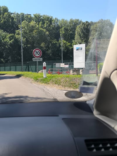 The image depicts a roadside scene in Saint-Pierre-de-Bœuf, France, viewed from inside a vehicle. Visible outside the vehicle are a speed limit sign indicating "70 RAPPEL" (reminder of a 70 km/h speed limit), a red and white striped bollard, and a large sign displaying project information near an active construction site. The sign appears to be related to the nearby "Z.A. DES GRANDS" industrial zone, as evidenced by the visible text "Z.A. DES GRANDS" and names of businesses or locations like "PNEUS BLAN", "MOLINA TRA", and "IMB BUS". A green fence runs along the perimeter, with trees forming a dense backdrop. The foreground is the dashboard of a vehicle with a registration sticker. The scene is bathed in daylight, under a clear sky.