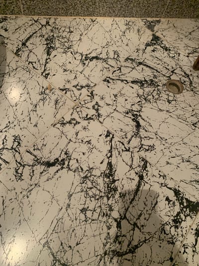 The image depicts a close-up view of a white tiled floor with black, marble-like veins. The tiles are arranged in a grid pattern, with visible grout lines. In the upper right quadrant of the image, a circular metal drain is present. The lighting suggests an indoor environment, likely a bathroom or similar wet area, with reflections visible on the glossy tile surface. There is a small, light-colored speck on the floor in the upper left quadrant, possibly debris. The image is taken from a slightly elevated perspective, looking down at the floor. The surrounding wall, visible in the upper edge of the frame, appears to be tiled with a different pattern, possibly a speckled grey. The specific location in Tabagrebi, Georgia, cannot be determined from the visual information provided in the image alone.