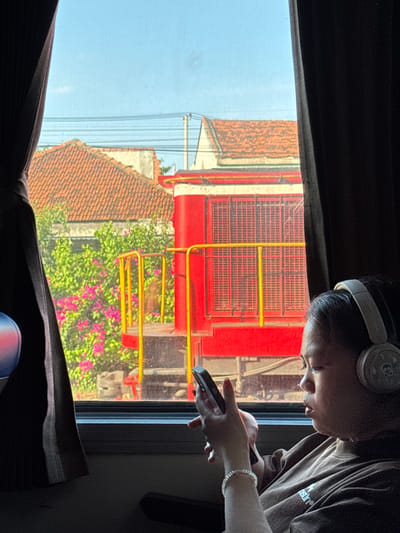 In Tuy Hoa, Vietnam, a young person wearing headphones and a brown t-shirt is looking at their phone. They have a bracelet on their left wrist. The person is seated inside, looking out a window. Outside the window, a red train is visible, with yellow railings. Behind the train, there are tiled roofs of houses and lush green foliage with pink flowers. Dark curtains frame the window on both sides. The sky is a clear blue.