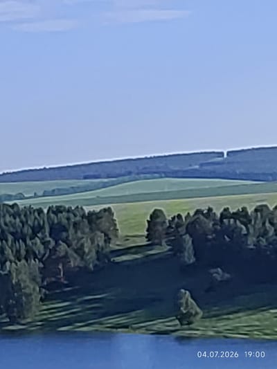 The image shows a wide-angle view of a rural landscape under a clear blue sky. In the foreground, a body of blue water occupies the lower portion of the frame. Beyond the water, a dense forest with dark green trees covers the lower middle ground, casting shadows on the grassy slopes behind it. Rolling green hills form the middle ground, with lighter green fields appearing as layered bands. A darker, forested ridgeline stretches across the horizon, with a visible gap or break in its continuity. The scene is illuminated by natural daylight, likely in the late afternoon given the timestamp in the bottom right corner which reads "04.07.2026 19:00". The location is identified as Konovalovo, Russia.