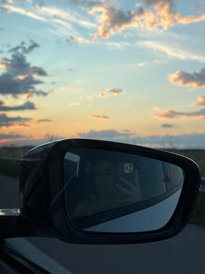The image shows a car's side mirror against a backdrop of a sky with clouds during sunset, likely taken while driving. The mirror reflects a person holding a smartphone, presumably taking the picture. The location, Opalchenets, Bulgaria, cannot be determined from the visual content.