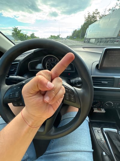 The image shows a first-person view from the driver's seat of a car on a highway. The driver's left hand, with the middle finger extended, is in the foreground, positioned over the steering wheel. The steering wheel has several control buttons. The dashboard displays include a speedometer, tachometer, and a central infotainment screen showing radio information. Outside the car, the view is of a highway with other vehicles, including a large truck, and trees lining the road. The sky is cloudy with patches of blue. The location is identified as the city of Severynivka, Ukraine.
