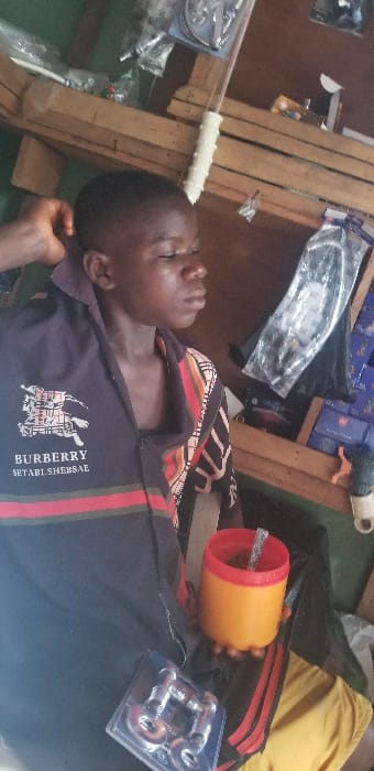 An image captured in Ado-Ekiti, Nigeria, depicts a young male individual seated indoors, likely within a shop or storage area. The individual, who has dark skin and short hair, is positioned facing right, with his eyes partially closed or downcast. A hand is visible touching the back of his neck or head on the left side of the frame. He is wearing a dark blue polo shirt with red and green stripes across the chest and a "BURBERRY SETABI.SHEBSAE" logo. A multi-patterned shirt with geometric shapes and stripes in black, white, red, and orange/brown is visible underneath or layered on his right side. In his right hand, he holds an orange and red thermos-style cup containing a clear straw. On a surface near his lap, a clear plastic blister pack containing two metal plumbing fixtures is visible. The background consists of wooden shelving units displaying various packaged merchandise, including clear plastic bags with components, blue cardboard boxes, and a hanging white ribbed rod. A dark green surface runs along the bottom edge of the shelving.