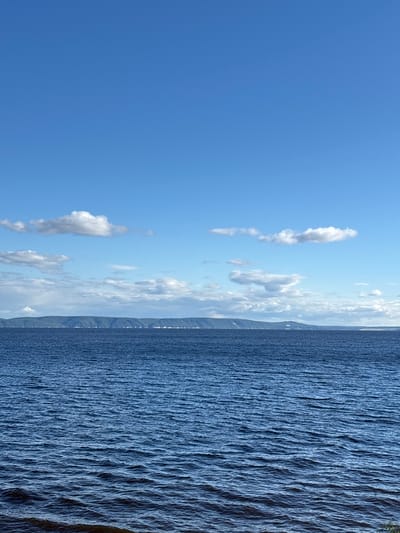 This media file shows a panoramic view of a body of water under a bright blue sky with scattered clouds. The scene is oriented vertically. On the right side of the image, the water is choppy, with small waves creating a textured surface. As the water moves towards the left, it becomes calmer, eventually appearing smooth and almost still in the far left portion of the frame. In the distance, along the horizon, there is a faint outline of mountains or hills, suggesting a landmass beyond the water. The sky is a clear, vibrant blue, with several fluffy white clouds present, particularly in the middle and upper sections of the image. The lighting indicates it is daytime, likely late morning or early afternoon, given the brightness. The overall impression is one of a serene natural landscape, possibly a lake or a calm sea. No people, objects, text, or specific activities are visible. The location context of Primorsky, Russia, suggests a coastal or lakeside setting within that region.