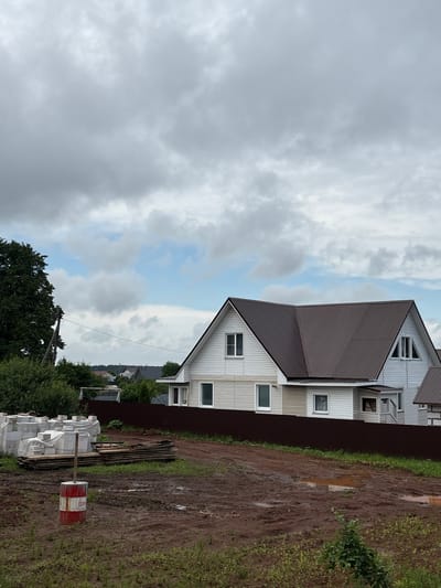 This image depicts a residential construction site in the city of Kstinino, Russia. In the foreground, a muddy area with puddles is visible, with a red and white painted barrel with a wooden pole sticking out of it. To the left, stacks of building materials, including wrapped white blocks and wooden planks, are present. A dark brown metal fence runs horizontally across the mid-ground. Behind the fence, a two-story white house with a brown metal roof is prominent, featuring several windows and white siding. Partially visible to the right is another house with a similar brown roof and white siding. The background shows a line of trees and other houses, indicating a suburban or rural setting. The sky is overcast with grey clouds, with patches of blue sky visible. The overall scene suggests ongoing construction and a damp, possibly post-rain environment.