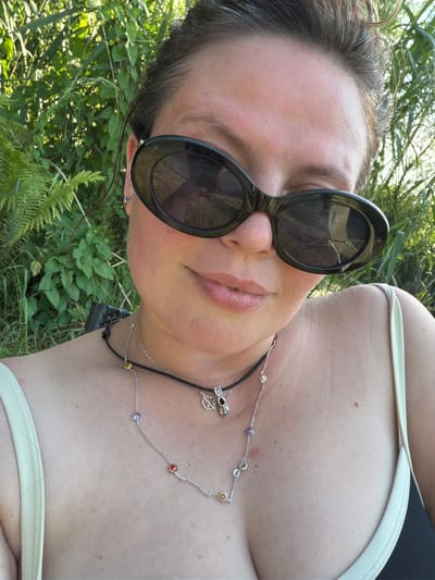 The media file shows a close-up, tilted shot of a person's face and upper body, outdoors. The subject is wearing large, dark-rimmed sunglasses and a light-colored strappy top. A silver necklace with colorful, small circular charms and a dark pendant is visible around their neck. The background consists of lush green foliage, suggesting a natural, possibly grassy or wooded, setting. Sunlight filters through the leaves, indicating it is daytime, likely with clear or partly cloudy weather. The person's head is tilted, and they appear to be looking upwards or to the side, with a relaxed or content expression suggested by their slightly parted lips. The overall impression is casual and relaxed, possibly taken during a summer day outdoors in Kamianka-Buzka, Ukraine. There is no visible text in the image.