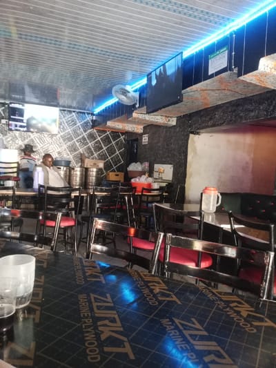 An interior scene of a commercial establishment, identified as being in Murang'a, Kenya, is captured. The space features a corrugated metal ceiling from which a blue LED strip light and a white oscillating fan are mounted. Walls exhibit varied textures, including dark patterned surfaces and a plain light section. Two flat-screen televisions are visible: the left screen displays a sports event, while the right screen shows a dark, indistinct image.

Furnishings include numerous black chairs with red cushioned seats and black tables. The table in the immediate foreground is covered with material labeled "ZURKT MARINE PLYWOOD," holding two clear plastic cups. In the mid-ground, two adult males are present; one stands partially obscured on the left, wearing a dark hat and light shirt, while another is seated to the right, facing away from the camera in a light shirt. Behind them, several metal beverage kegs are stacked, and a counter area contains multiple plastic containers filled with ice and a beverage dispenser. A clear plastic jug with an orange lid is placed on a table in the mid-ground right. A white sign affixed to the upper right wall displays "LIPA NA M-PESA Till No: 3171026," with a smaller sign below it stating "TRY AS YOU ORDER." No active interactions are depicted between individuals or with objects.