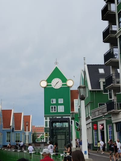 The image shows a street scene in Zaandam, Netherlands, with several distinctive green buildings. In the center, a tall green building features a large clock on its facade and a white circular element to its left, possibly another clock face or decorative element. This building has multiple windows and a pointed roof. To the right, another green building with a dark tiled roof is visible, also with windows and balconies. In the foreground, a street or walkway is lined with a bright green fence. Several people are walking or standing on this walkway. To the left of the central green building, there are smaller, brightly colored buildings with red tiled roofs. One of these buildings is predominantly blue with white trim. The sky is overcast with a pale gray cloud cover. The overall impression is of a well-maintained urban environment with a unique architectural style.